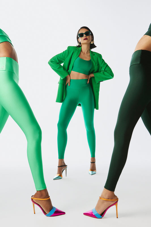 Woman in a bold green Glossy leggings set with a blazer and sunglasses, hands on hips for a retro-inspired portrait